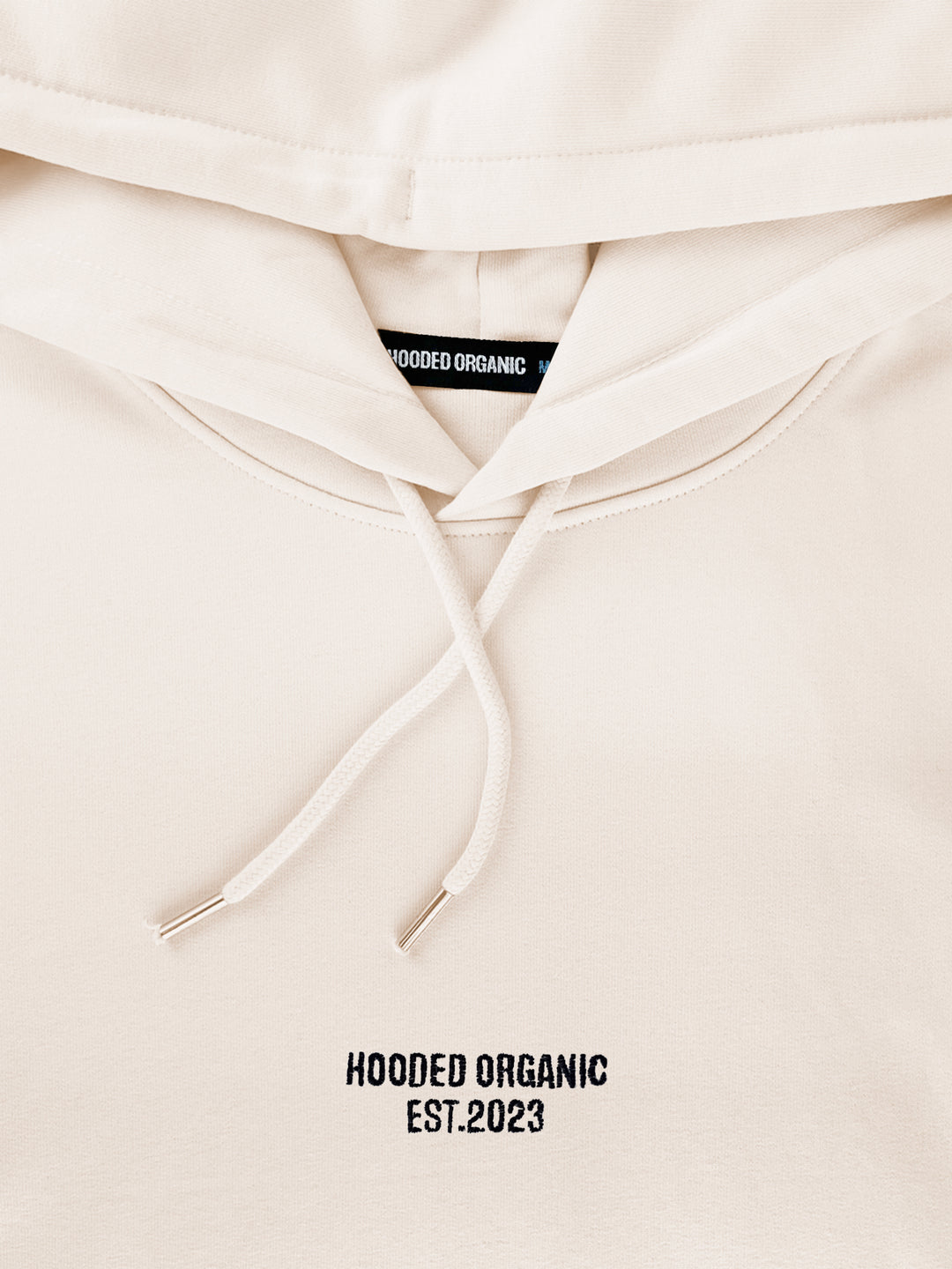 Nærbillede af Hooded Organic-logo på beige hoodie – lavet i 100% økologisk bomuld
