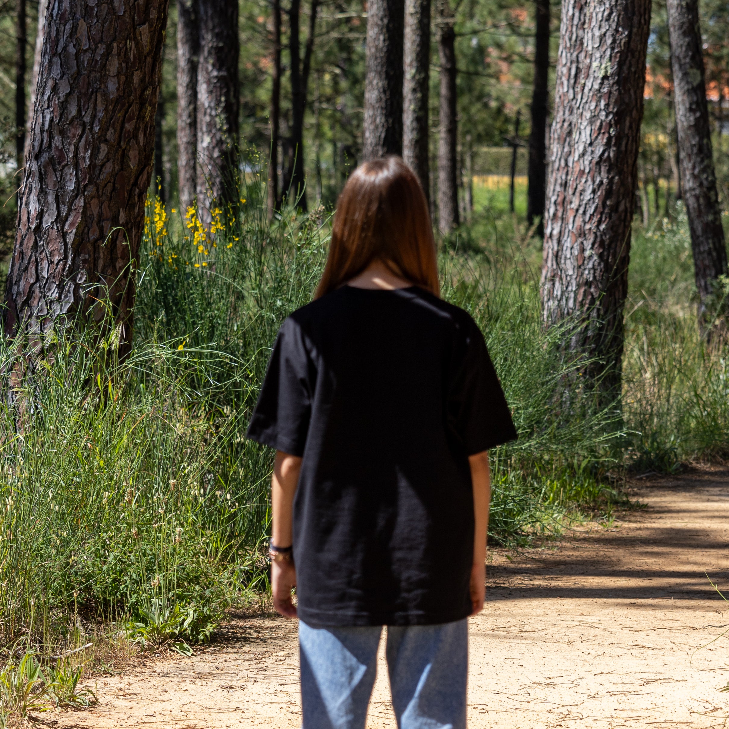 Kvindelig model i sort T-shirt med ryggen til i skoven – Hooded Organic-logo på fronten og økologisk bomuld